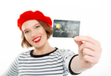 smiling-ginger-woman-showing-credit-card-camera-grey-focus-card